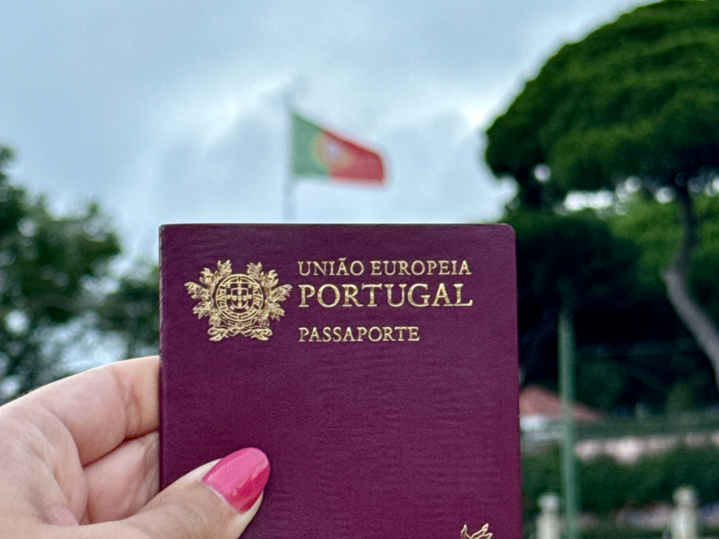 Mão segurando um passaporte português vermelho em primeiro plano, com a bandeira de Portugal hasteada e árvores ao fundo sob céu azul.